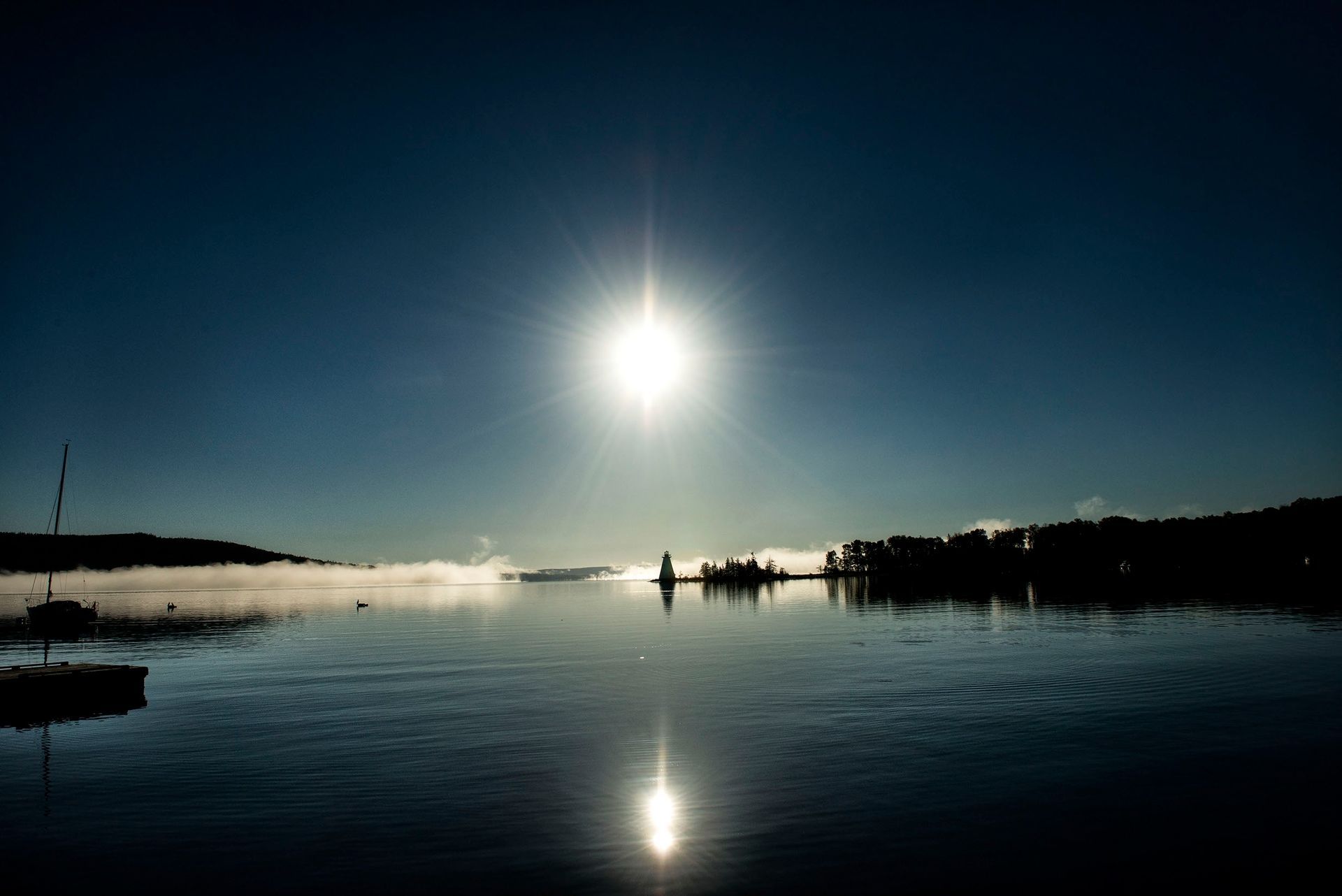 Een felle zon boven kalm water bij zonsopgang, met silhouetten van boten en de kustlijn die in het wateroppervlak worden weerspiegeld.