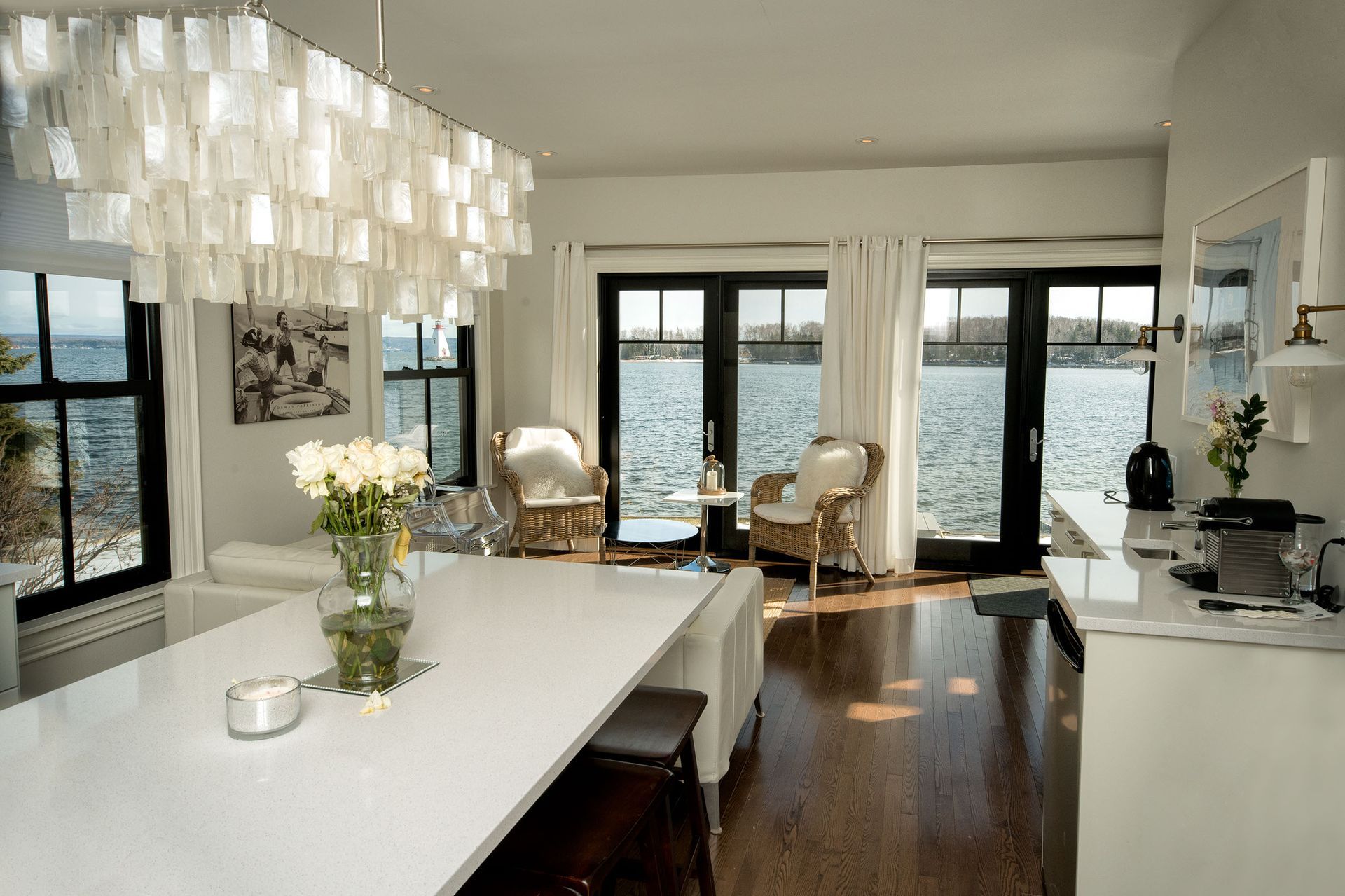 Lichte eetkamer met grote ramen, witte tafel, moderne kroonluchter en uitzicht op het water.