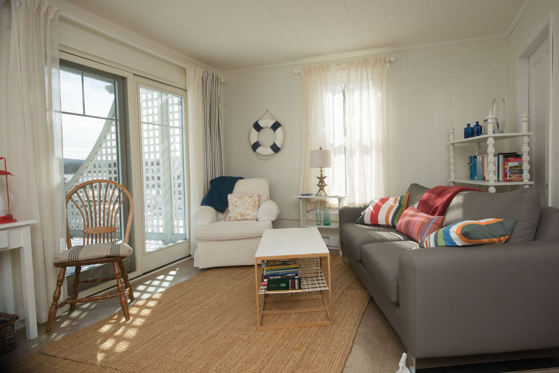 Lichte woonkamer met een grijze bank, witte fauteuil, rieten stoel en openslaande deuren die veel zonlicht binnenlaten.