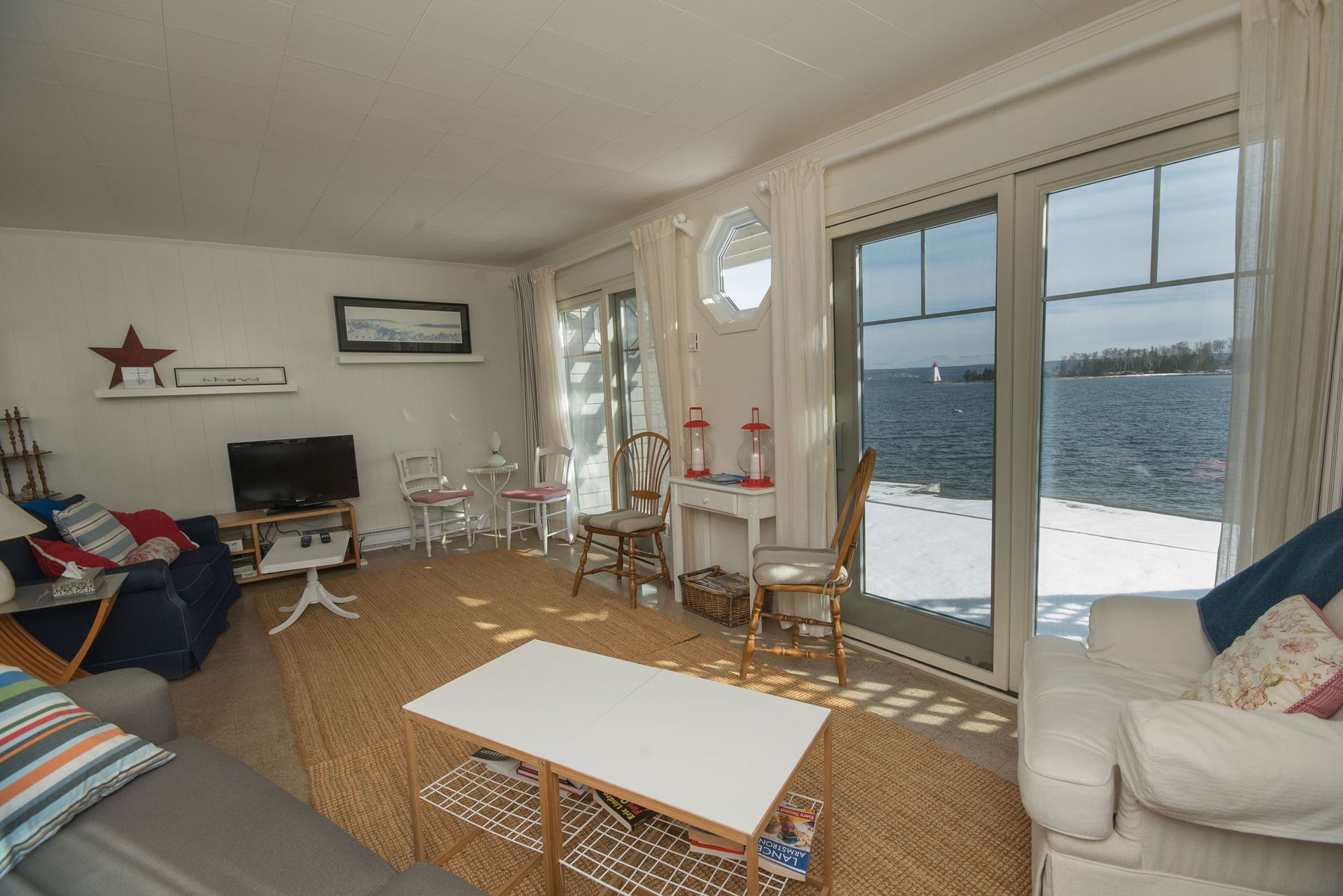 Lichte woonkamer met grote ramen die uitkijken op het water, witte meubels, een vloerkleed met patroon en een tv.