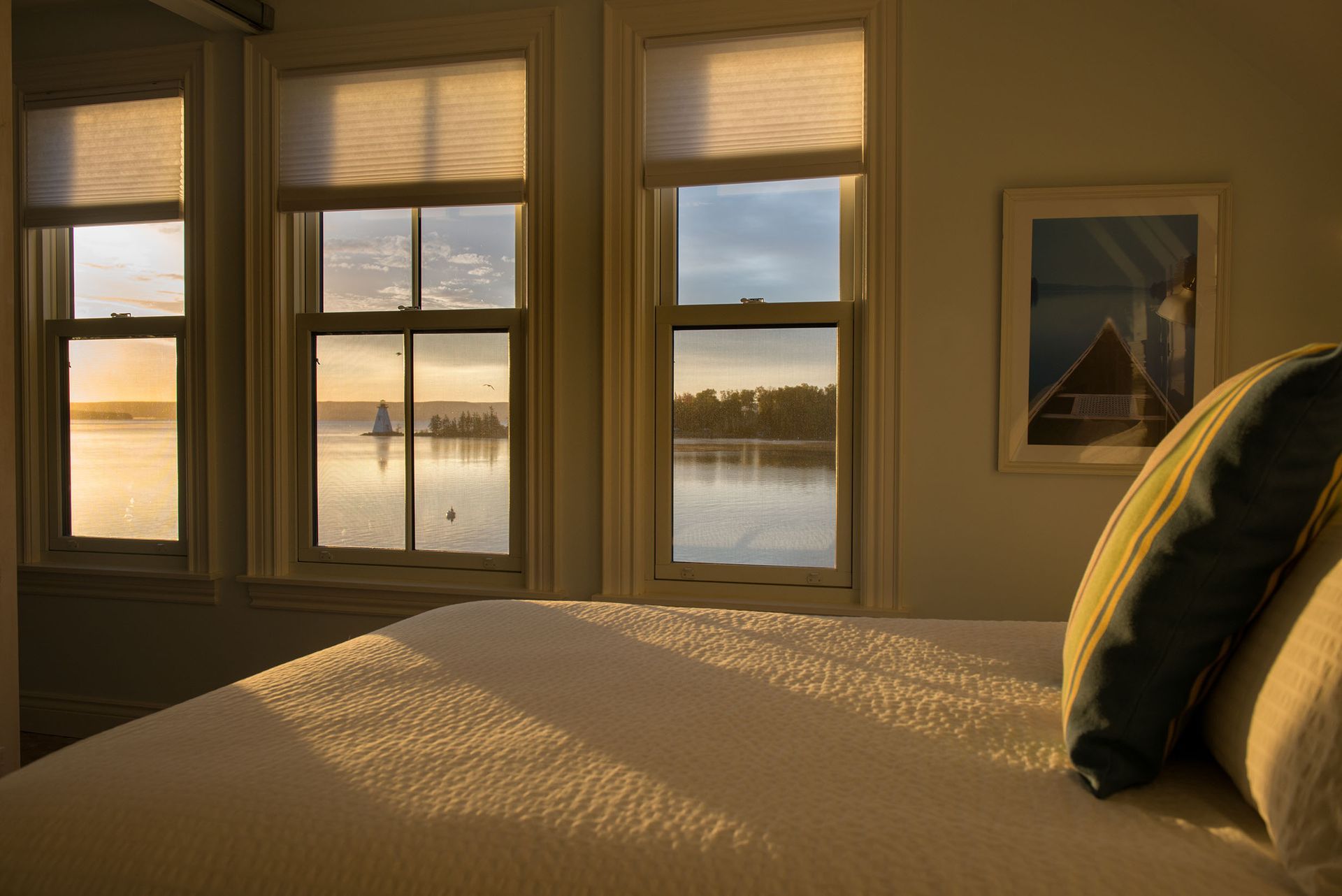 Zonnige slaapkamer met bed bij drie ramen die uitkijken op een rustig wateroppervlak bij zonsondergang.