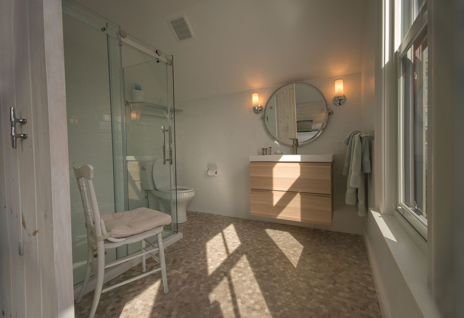 Zonnige badkamer met stoel, ronde spiegel, dubbele wastafel, douche en tegelvloer.