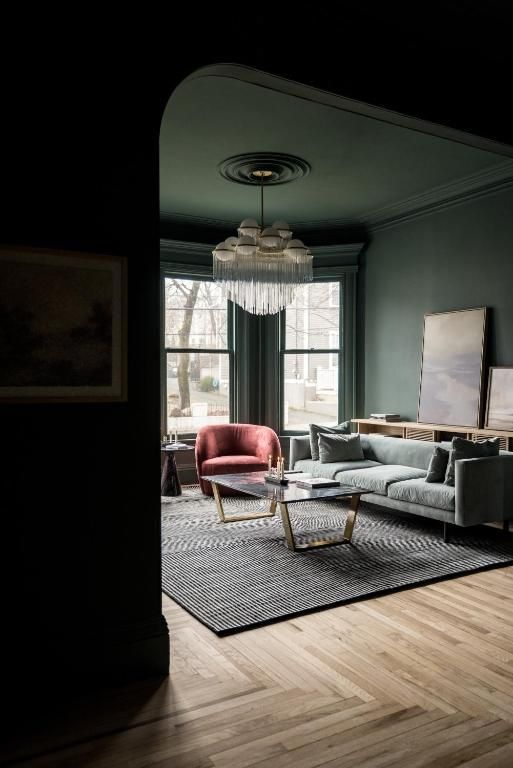 Elegante woonkamer met een grijze bank, rode fauteuil, kroonluchter, groot raam en houten vloer in visgraatpatroon.