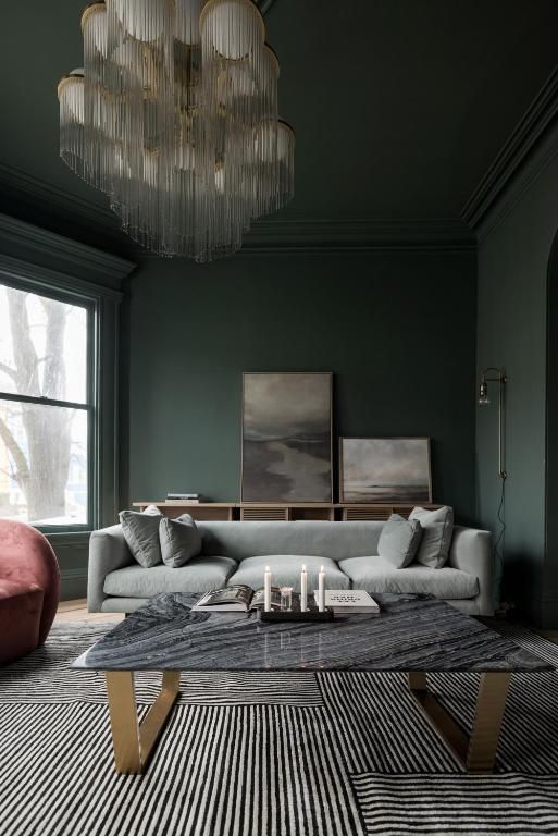 Moderne donkergroene woonkamer met een grijze bank, een vloerkleed met patroon, een glazen salontafel en een grote kroonluchter.