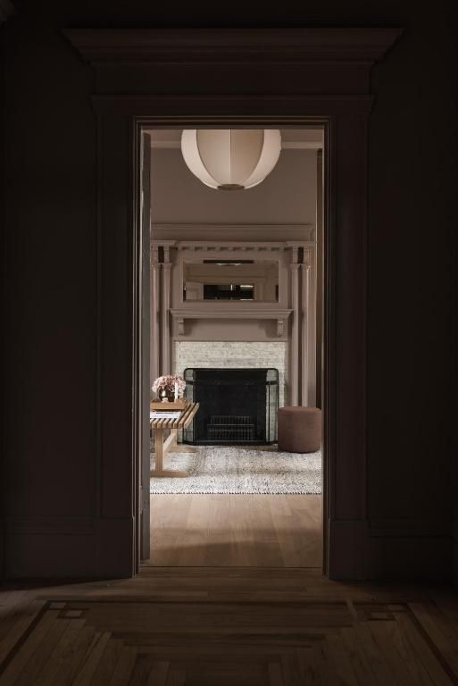 Een schemerige deuropening omlijst een lichte woonkamer met een open haard, een ronde hanglamp en een fauteuil.