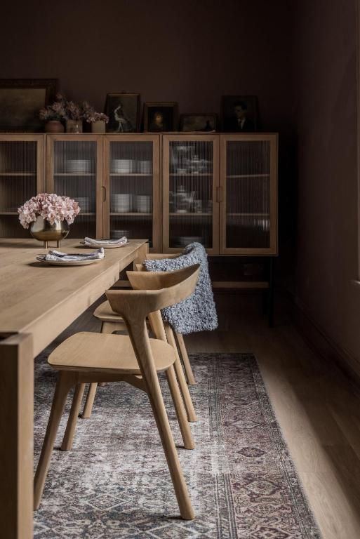 Eetkamer met houten tafel, gevlochten stoelen, een vloerkleed met patroon en een vitrinekast in zacht licht.
