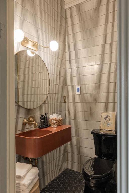 Kleine betegelde badkamer met koperen wastafel, ronde spiegel, wandlampen en een zwart toilet.