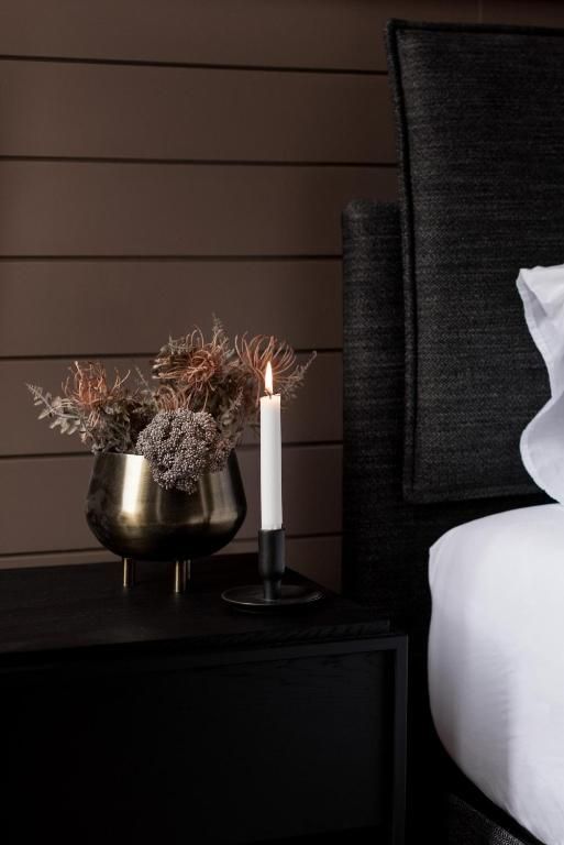 Modern nachtkastje voor de slaapkamer met een messing vaas, gedroogde bloemen, een brandende kaars en wit beddengoed naast een donker gestoffeerd hoofdbord.
