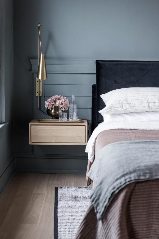 Moderne slaapkamer met donkerblauwe muren, zwevend nachtkastje, bloemen, lamp en een bed met wit en grijs beddengoed.
