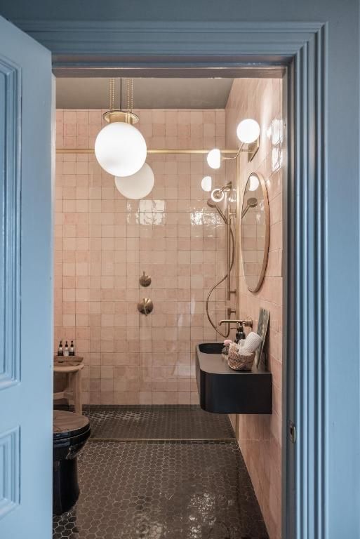 Kleine badkamer met roze tegels, blauwe deur, ronde hanglampen, spiegel, wastafel en douche.