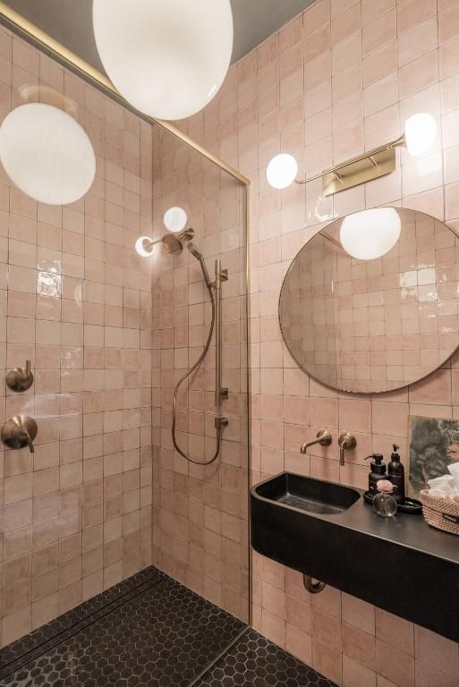 Badkamer met roze tegels, ronde spiegel, zwarte wastafel, douche en bolvormige lampen.