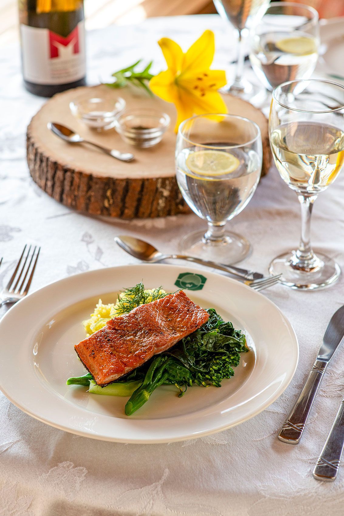 Zalmfilet met groenten op een wit gedekte tafel met wijnglazen en een geel bloemstuk als tafeldecoratie.
