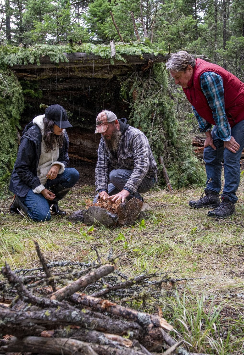 Drie mensen hurken in een open plek in het bos en onderzoeken een boomstam naast een stapel takken.