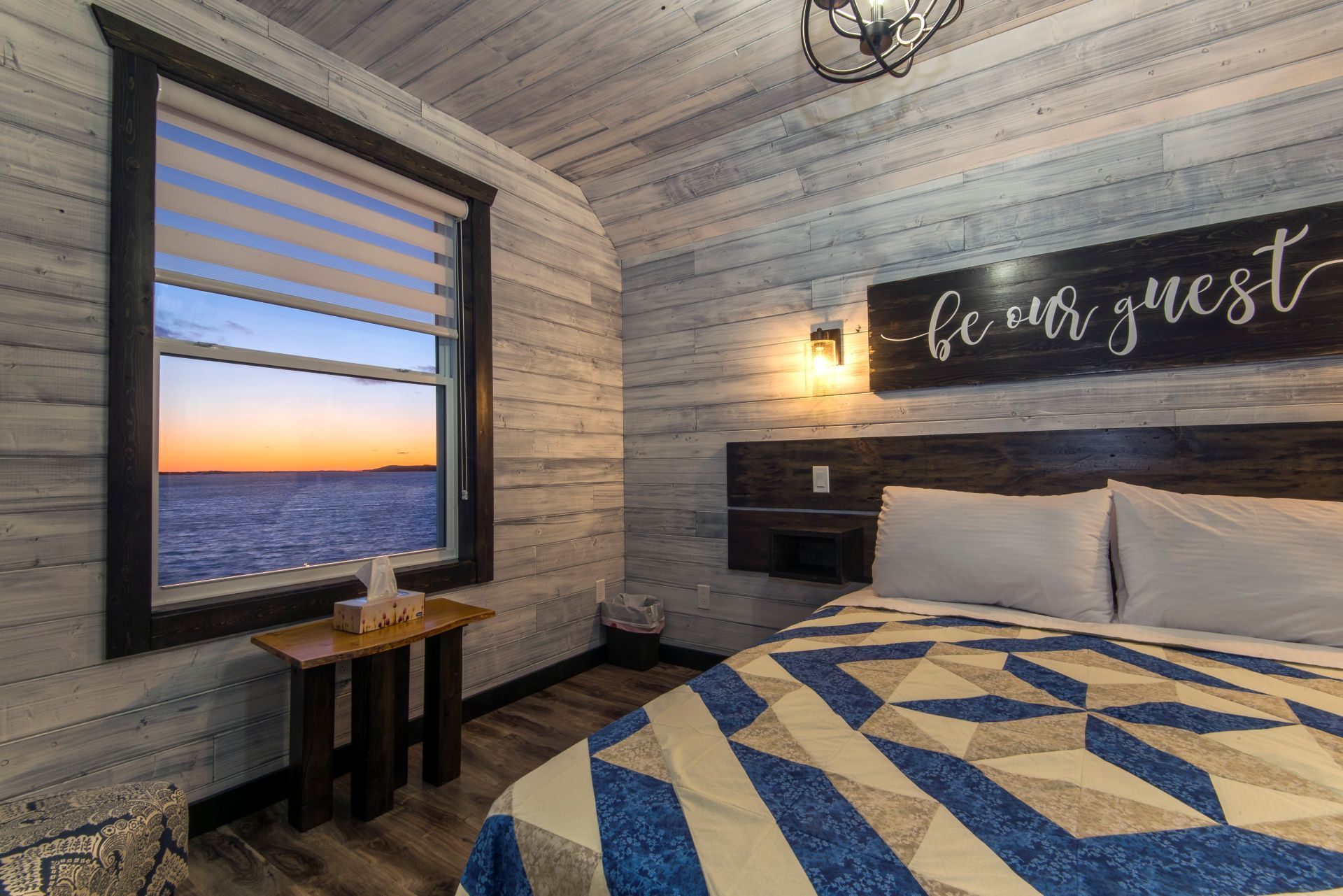 Gezellige slaapkamer in een blokhut van Escape by the Sea in Newfoundland met een bed, een raam met uitzicht op de oceaan en een bordje met 'wees onze gast' aan de muur.