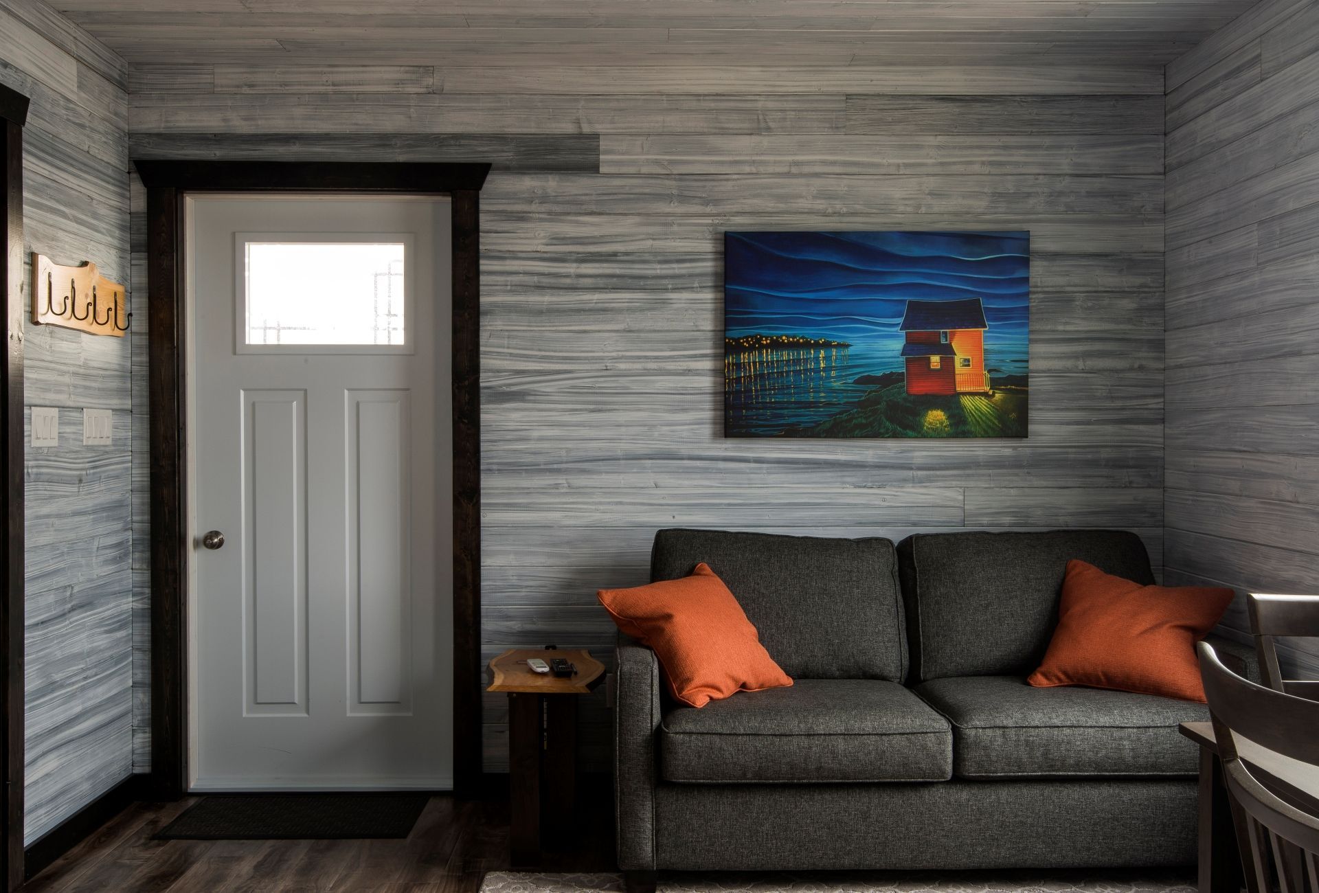 Grijze woonkamer in een huis van van Escape by the Sea in Newfoundland met witte deur, donkere bank, oranje kussens en een kleurrijk schilderij.