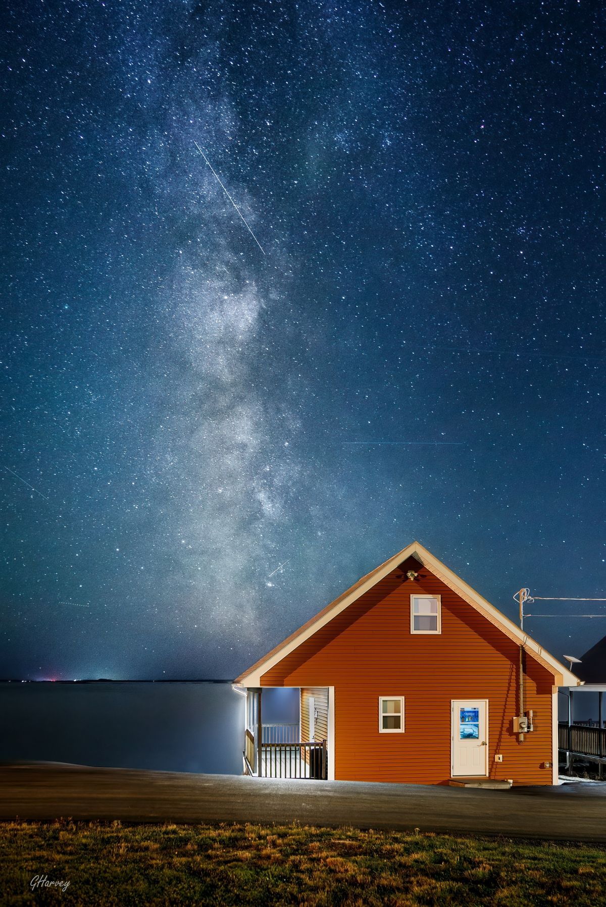 Rood huis van Escape by the Sea in Newfoundland aan een rustige zee onder een sterrenhemel met de Melkweg zichtbaar.