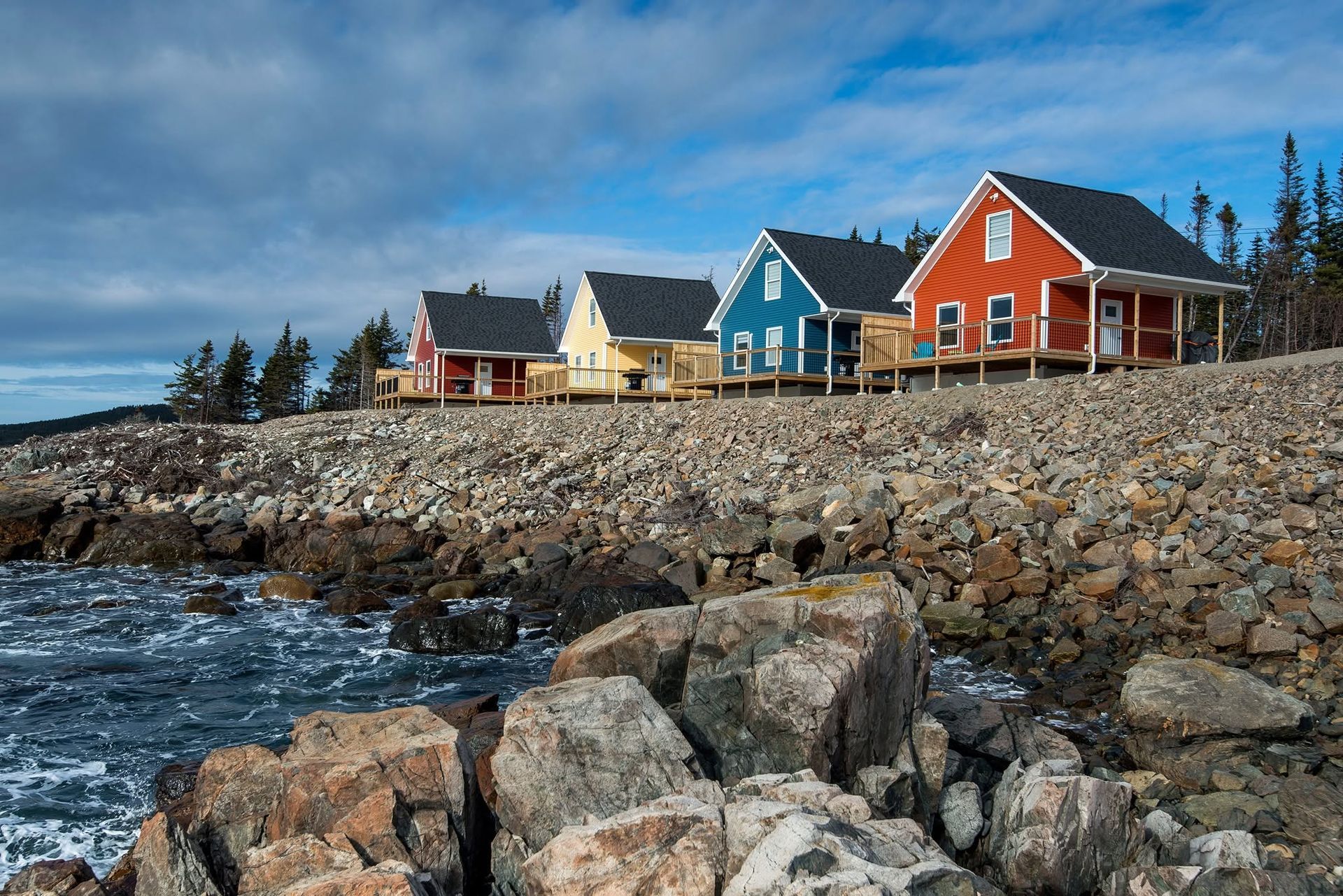 Kleurrijke huisjes van Escape by the Sea in Newfoundland aan zee op een rotsachtige kustlijn, naast de ruige oceaan onder een blauwe hemel.