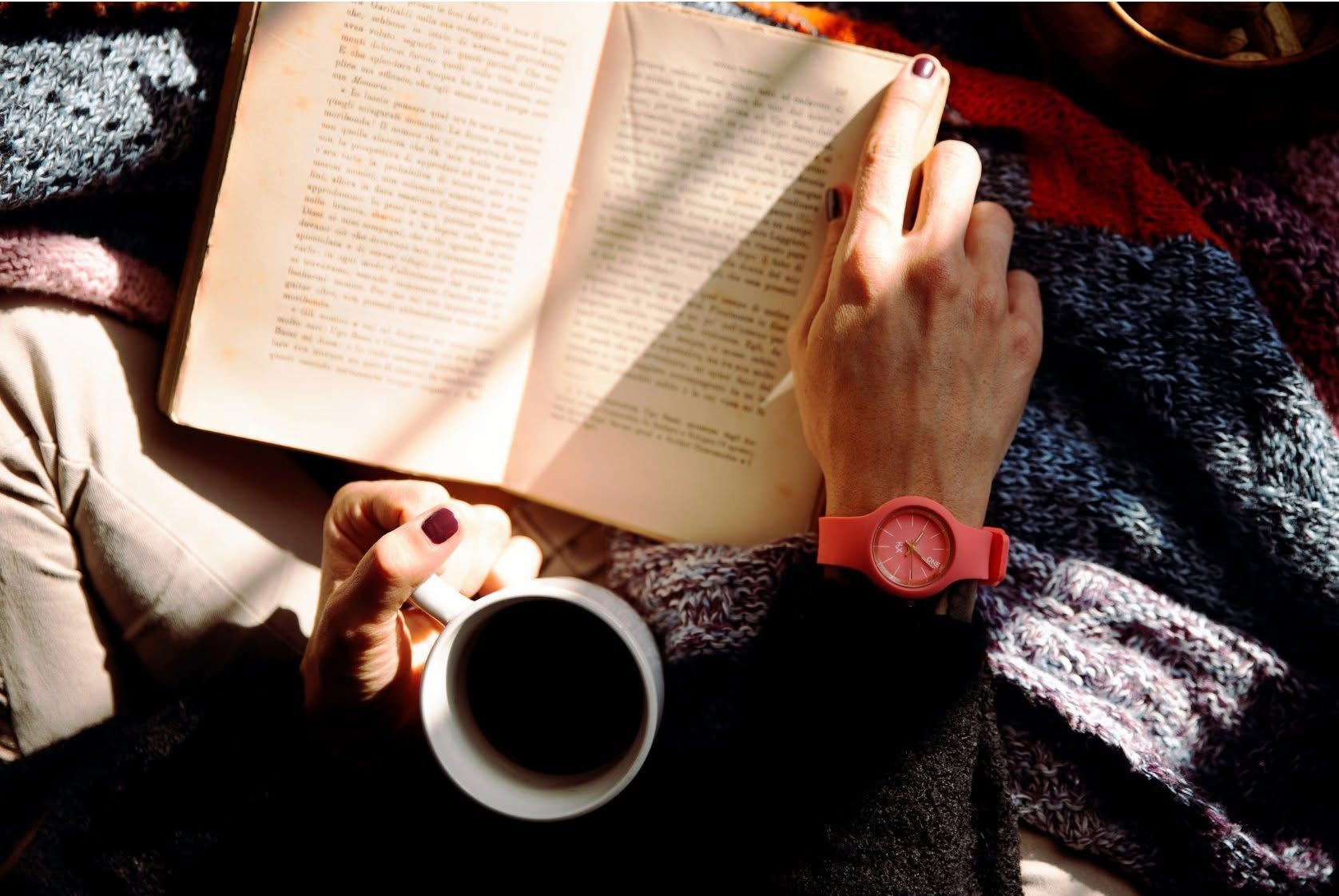 Een persoon leest een open boek naast een kop koffie, met een rood horloge om zijn pols, liggend op een deken.