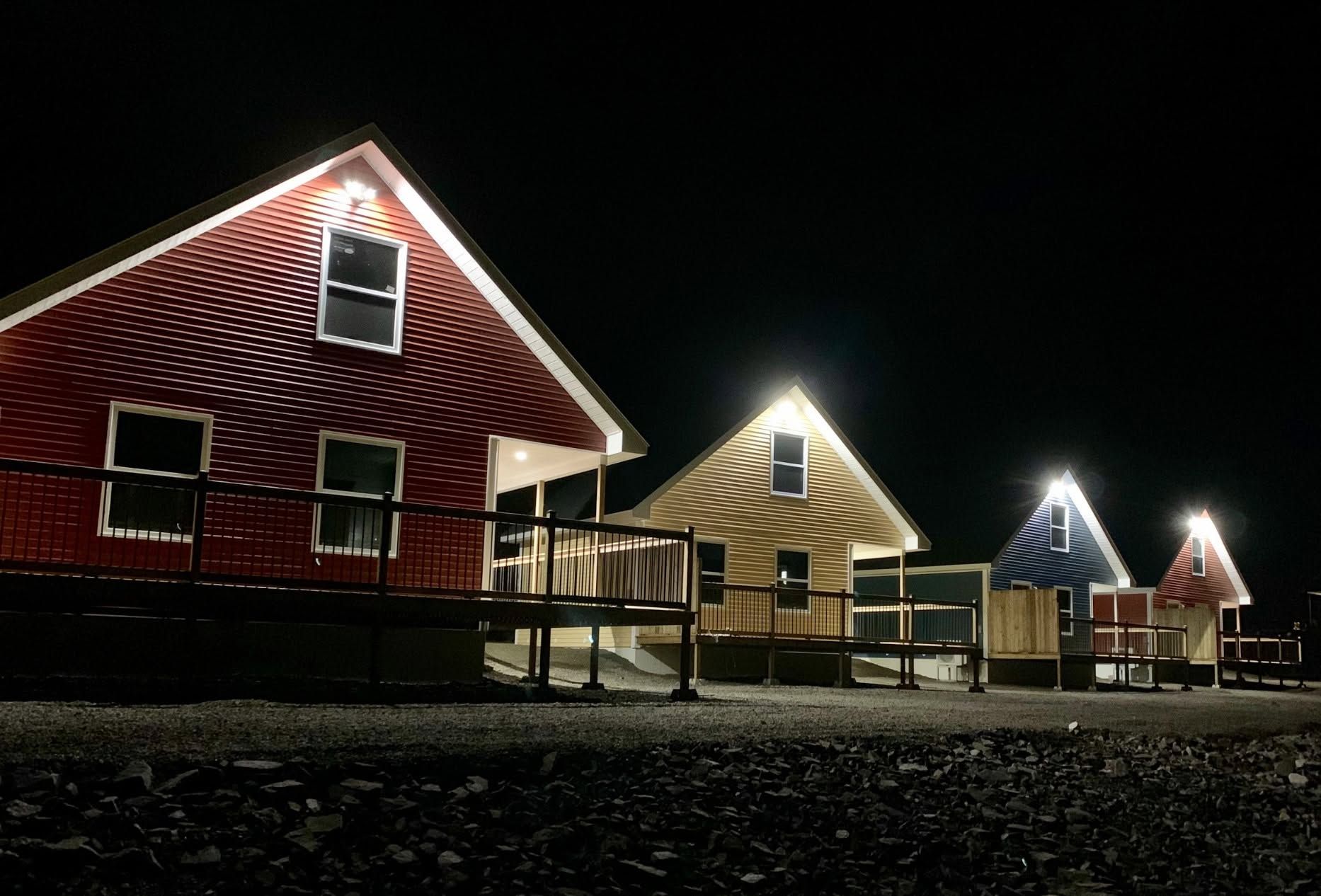 Een rij kleine, verlichte huizen van Escape by the Sea in Newfoundland 's nachts met rode, beige en blauwe gevels op een donkere heuvel.
