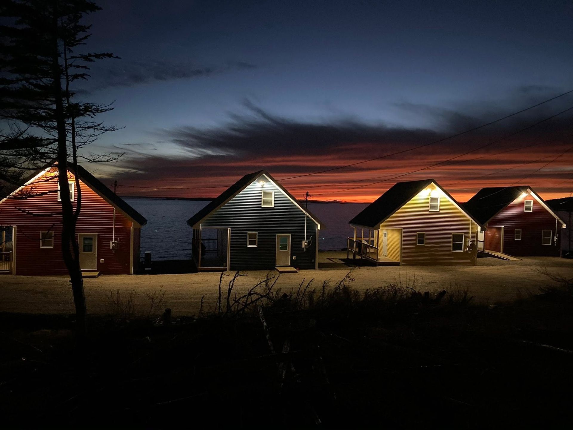 Een rij kleine, verlichte houten huizen van Escape by the Sea in Newfoundland in de schemering onder een donkere hemel met een rood-oranje zonsondergang.
