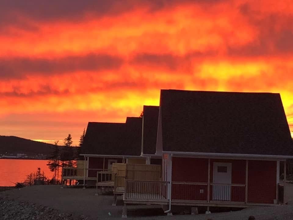 Hutten aan de zee van Escape by the Sea in Newfoundland bij zonsondergang, met een levendige oranje-rode lucht die in het water weerspiegelt.