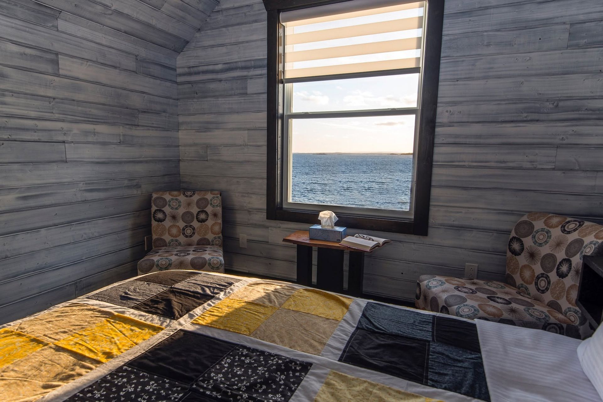 Gezellige slaapkamer met twee bedden met een patroon, een raam met uitzicht op zee en grijze houten lambrisering.