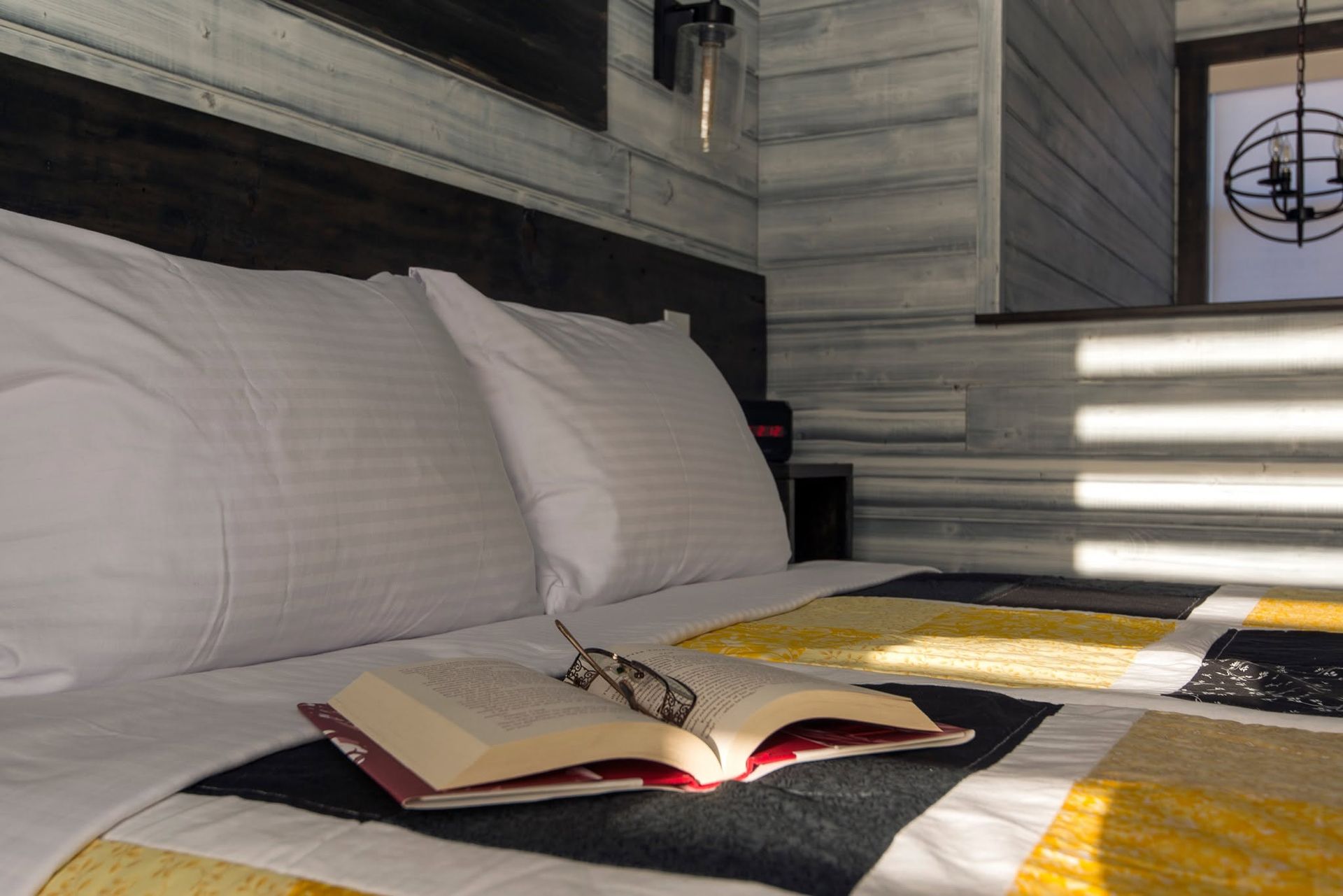 Een open boek op een bed met witte kussens en een zwart, geel en grijs dekbed in een moderne slaapkamer.