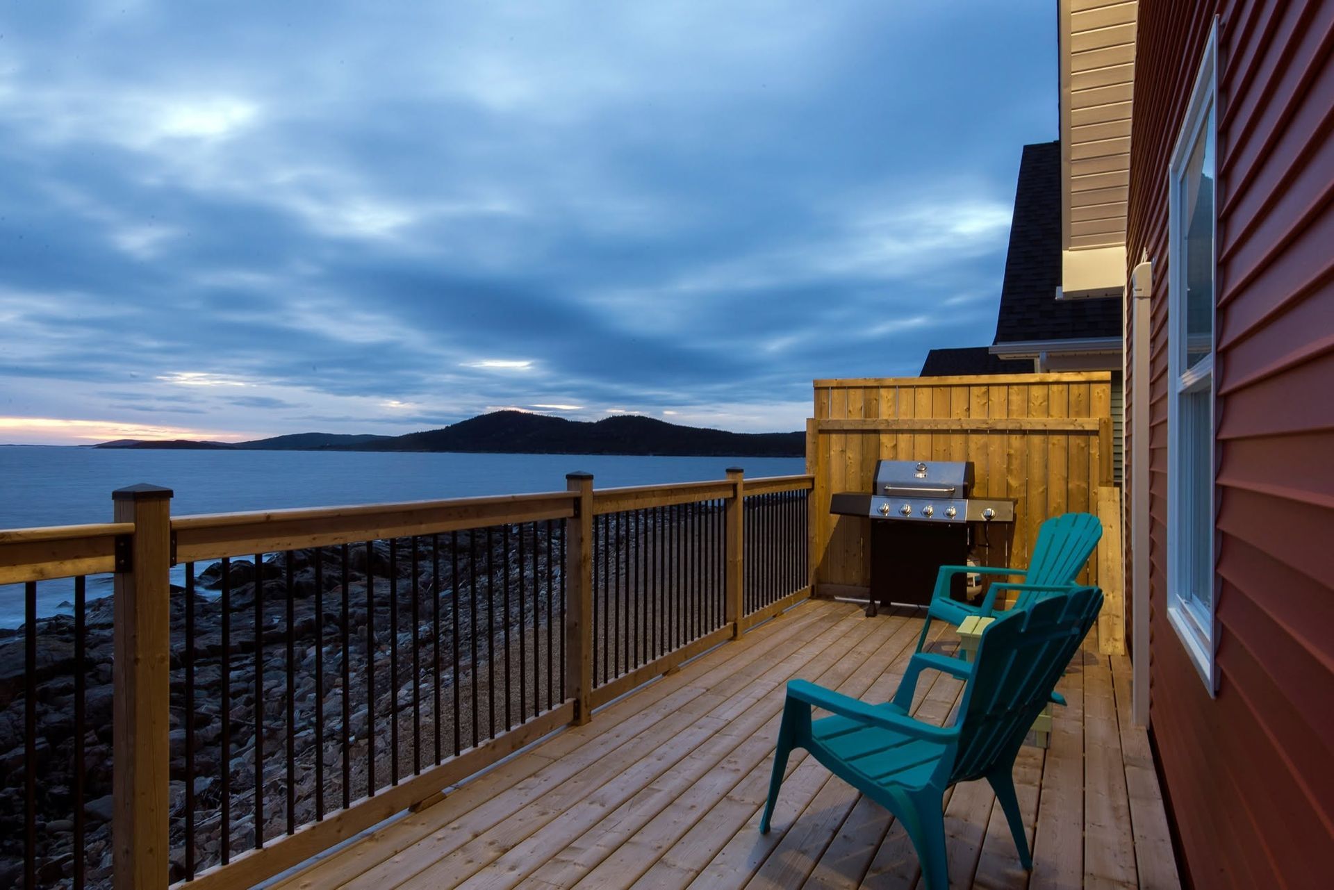 Houten terras met turquoise stoelen, met uitzicht op een rotsachtige kustlijn bij zonsondergang.