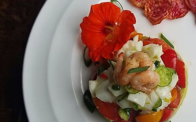 Salade met garnalen, komkommer, rode tomaat en een rode bloemgarnering op een wit bord.