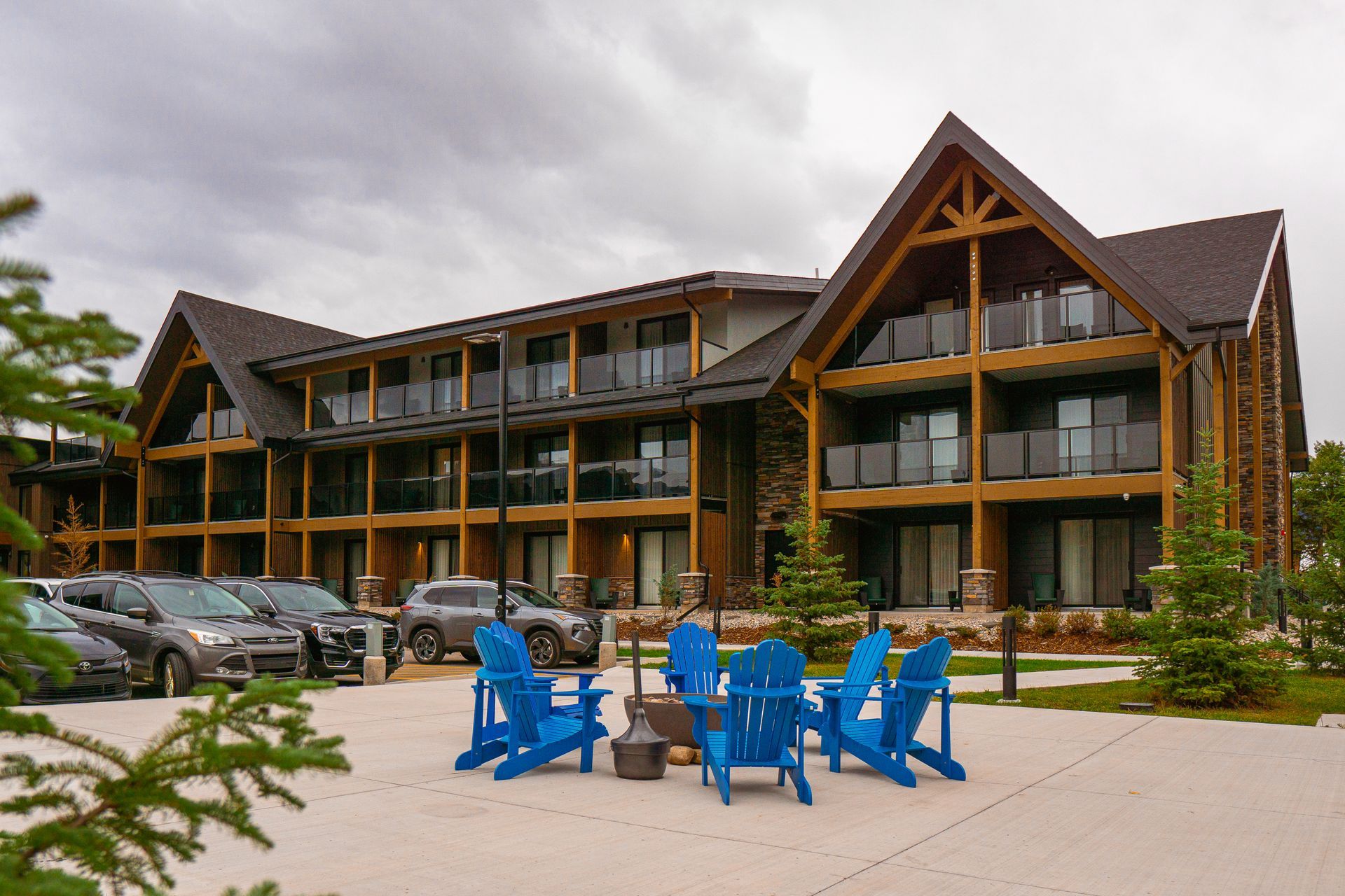Het hoofdgebouw van Forest Park Hotel Jasper met houten balken, geparkeerde auto's en blauwe stoelen op een betegeld terras onder een bewolkte hemel.