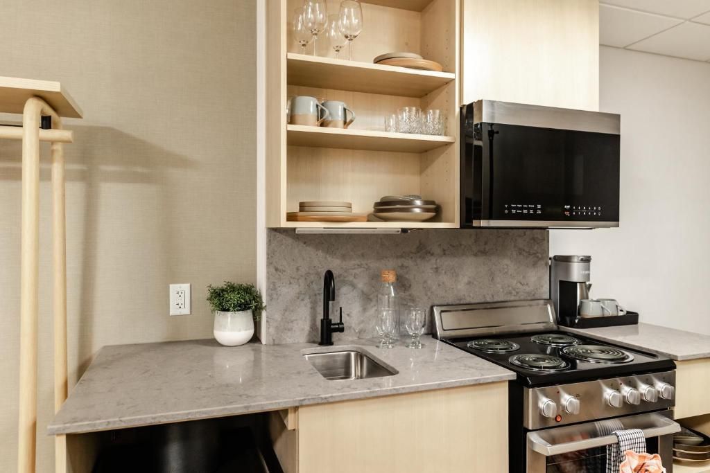 Compacte, moderne kitchenette van Forest Park Hotel, Jasper met lichte houten kasten, marmeren aanrechtblad, zwart fornuis en open planken.