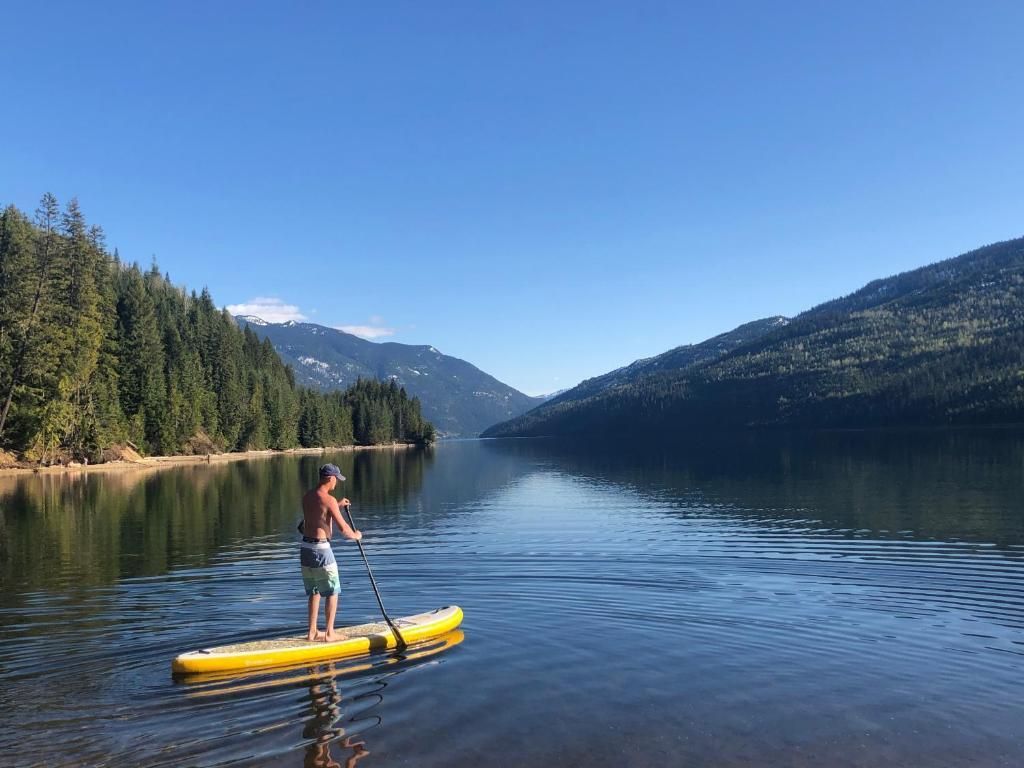 Een persoon staat op een geel paddleboard op een kalm meer, omgeven door beboste bergen.