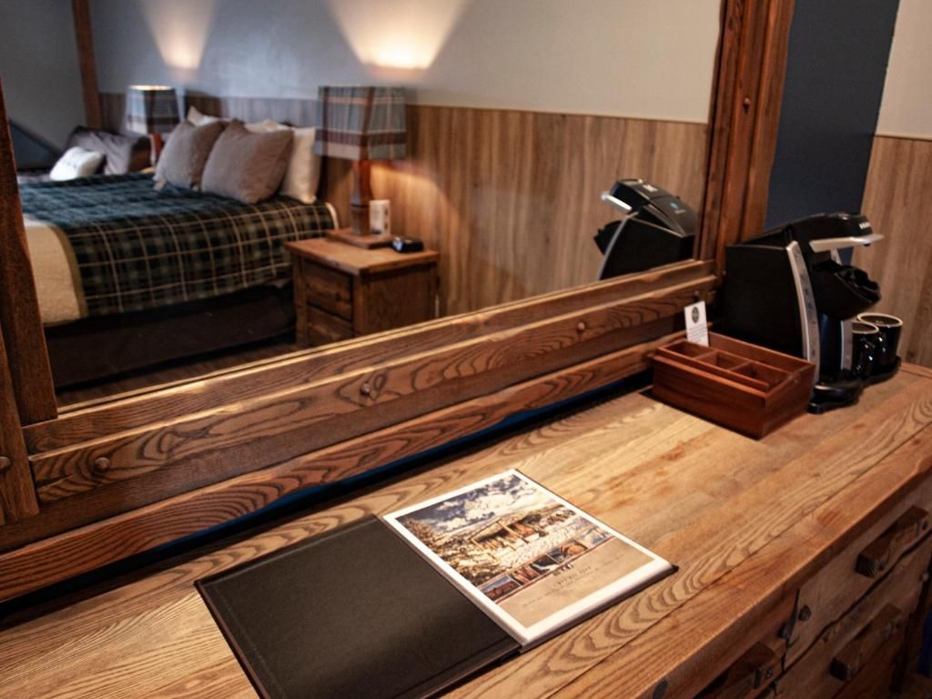 Hotelkamerbureau met spiegel, zwart notitieboekje, brochure, lamp en twee koffiezetapparaten op een houten oppervlak.