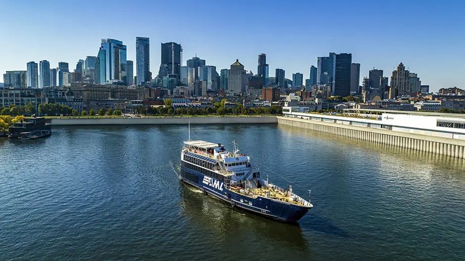 Een bootje op een kalme rivier met de skyline van een stad en gebouwen aan de waterkant op de achtergrond, onder een helderblauwe hemel.