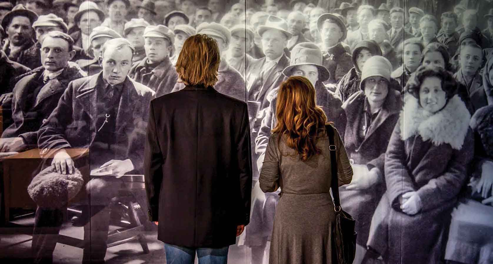Twee mensen staan ​​voor een grote zwart-wit muurschildering van een menigte in een galerie.