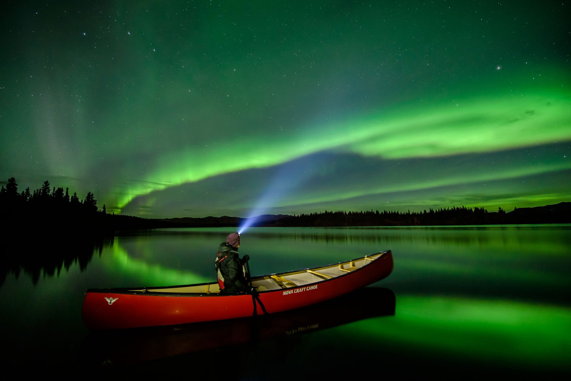 @hynesightadventures Persoon in een rode kano in Yukon op een rustig meer onder het groene noorderlicht, 's nachts