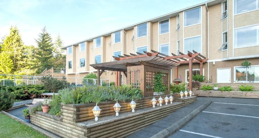De buitenkant van een beige rijtjeshuis met een aangelegde tuin, pergola en parkeerplaats ervoor.