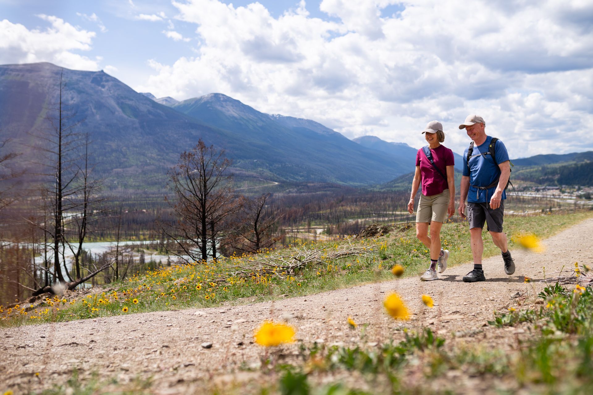 Twee wandelaars nabij Jasper National Park lopen over een bergpad met wilde bloemen en een meer beneden, onder een bewolkte hemel.