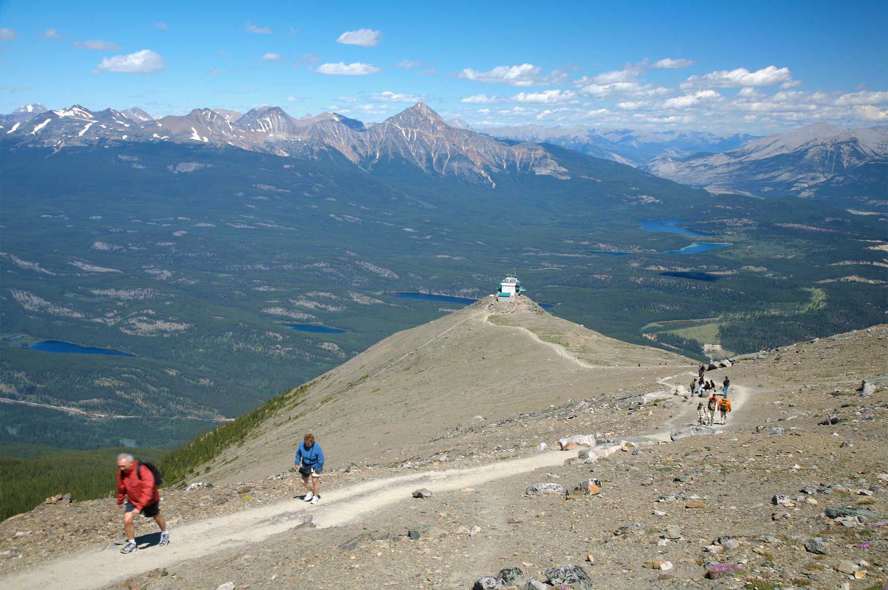 Wandelaars op een rotsachtig bergpad in Jasper National Park met uitzicht op een brede vallei en verre, met sneeuw bedekte bergtoppen.