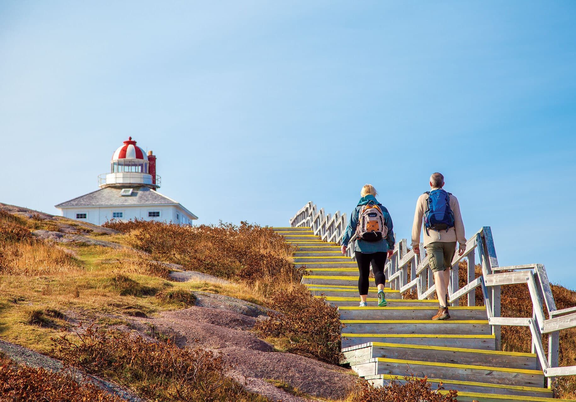 Twee wandelaars beklimmen trappen richting een rood-witte vuurtoren op een zonnige heuvel aan de kust.