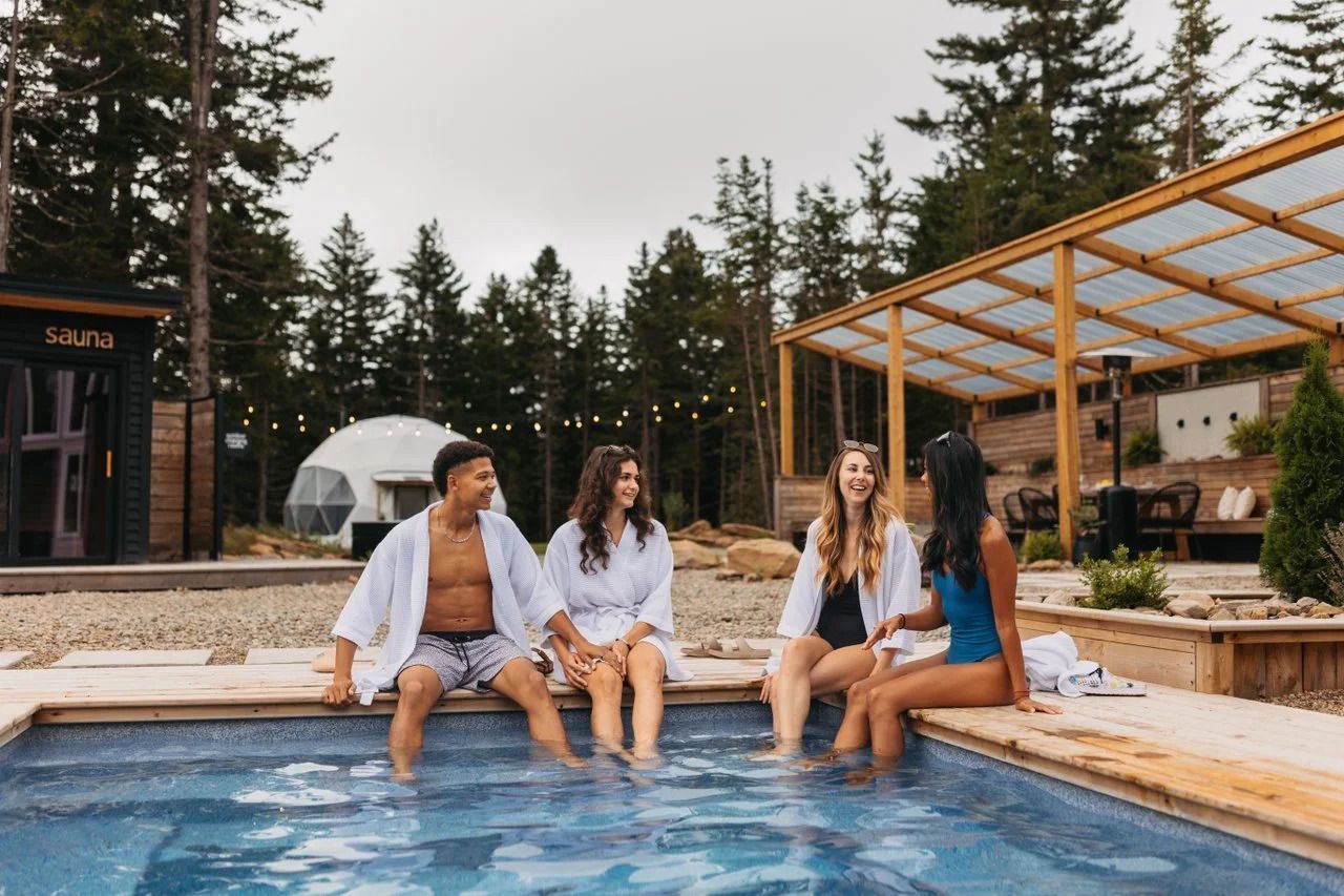 Vier vrienden zitten lachend op de rand van een zwembad in een moderne buitenjacuzzi met een houten overkapping en bomen op de achtergrond.
