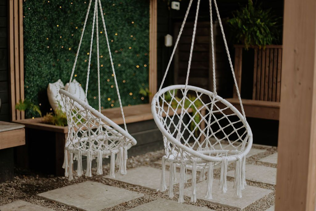 Twee witte macramé hangstoelen op een overdekt terras met groene wanddecoratie en een vloerkleed met patroon.