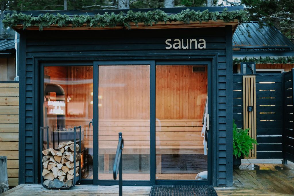 De buitenkant van een kleine, donkerblauwe sauna met glazen deuren en opgestapeld brandhout binnenin.