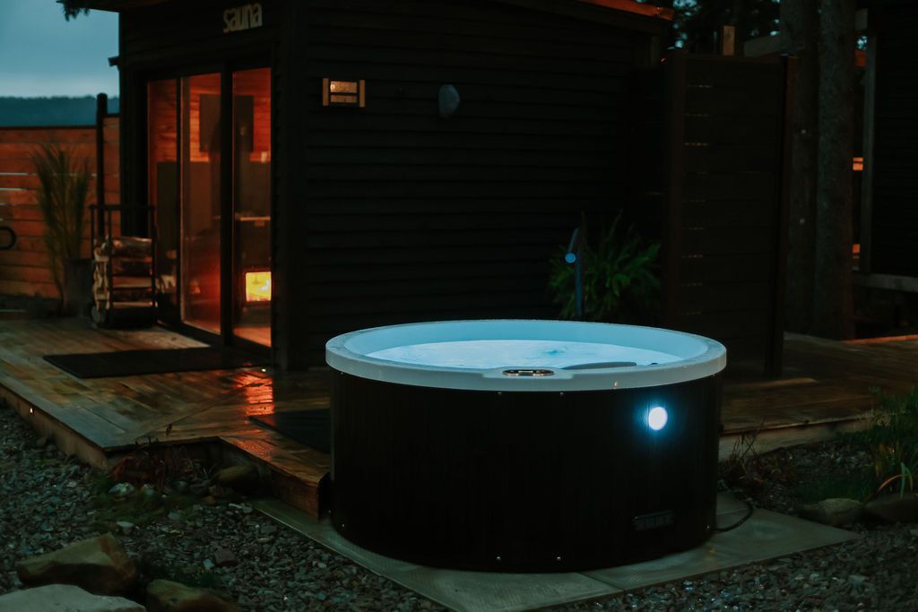 Een kleine, ronde, blauw oplichtende buitenjacuzzi staat 's avonds naast een donkere hut.