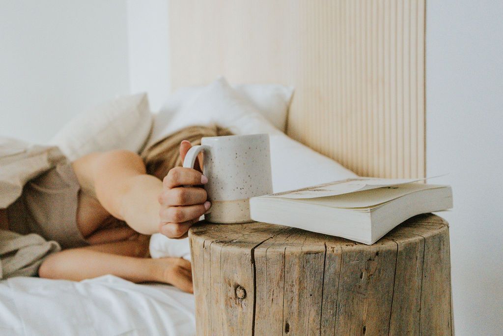 Persoon met een mok naast een stapel boeken op een rustieke houten boomstronk in een knus bed.
