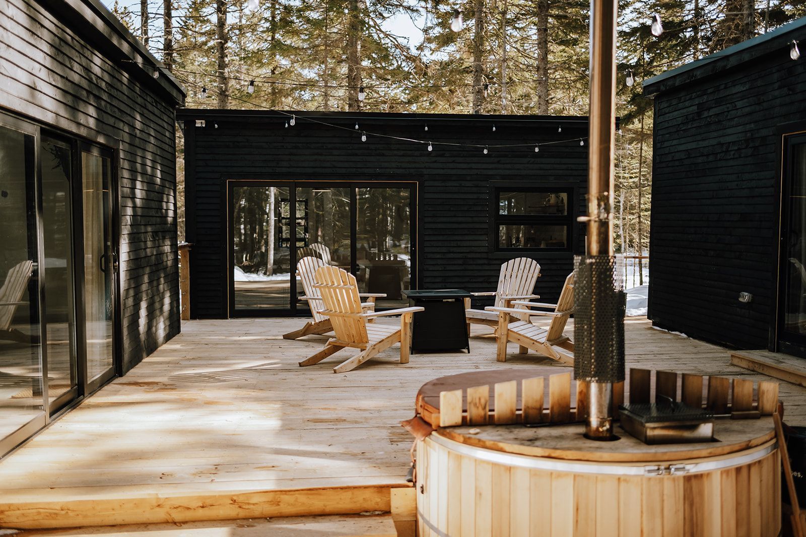 Modern terras bij een blokhut met ligstoelen, een vuurplaats en een jacuzzi te midden van hoge dennenbomen.