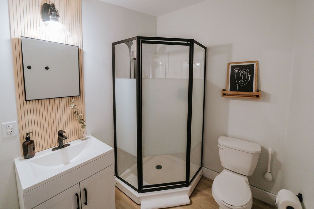 Badkamer met een douchecabine met zwart frame, een witte wastafel, een toilet en een klein wandplankje in een lichte hoek.