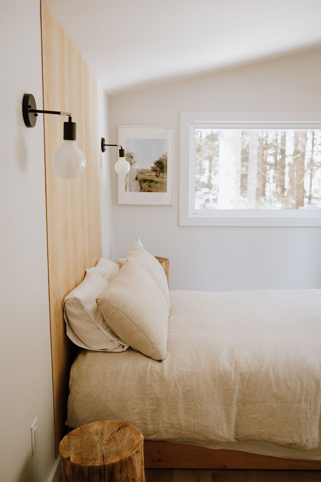 Gezellige slaapkamer met beige beddengoed, een houten accentmuur, een nachtlampje en een raam met uitzicht op besneeuwde bomen.