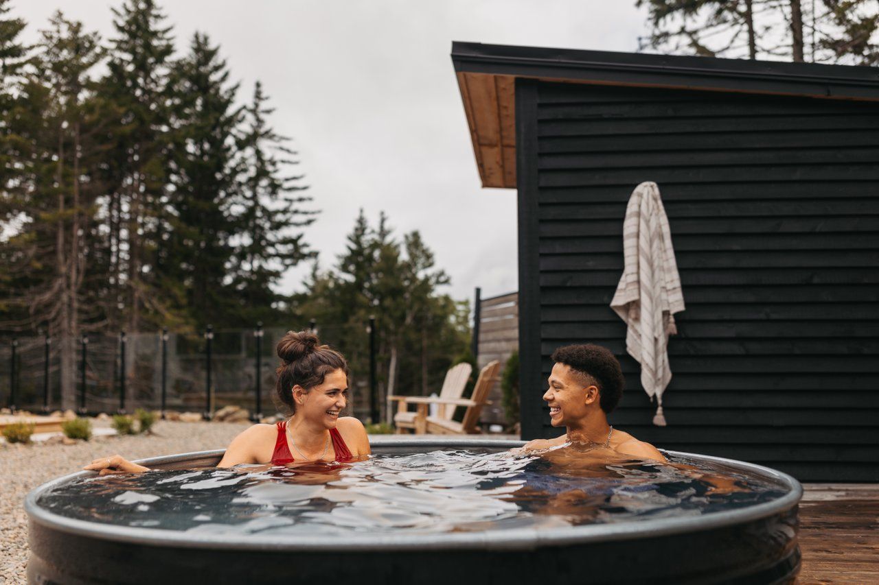 Twee mensen ontspannen in een buitenjacuzzi naast een donkergroene blokhut en bomen.