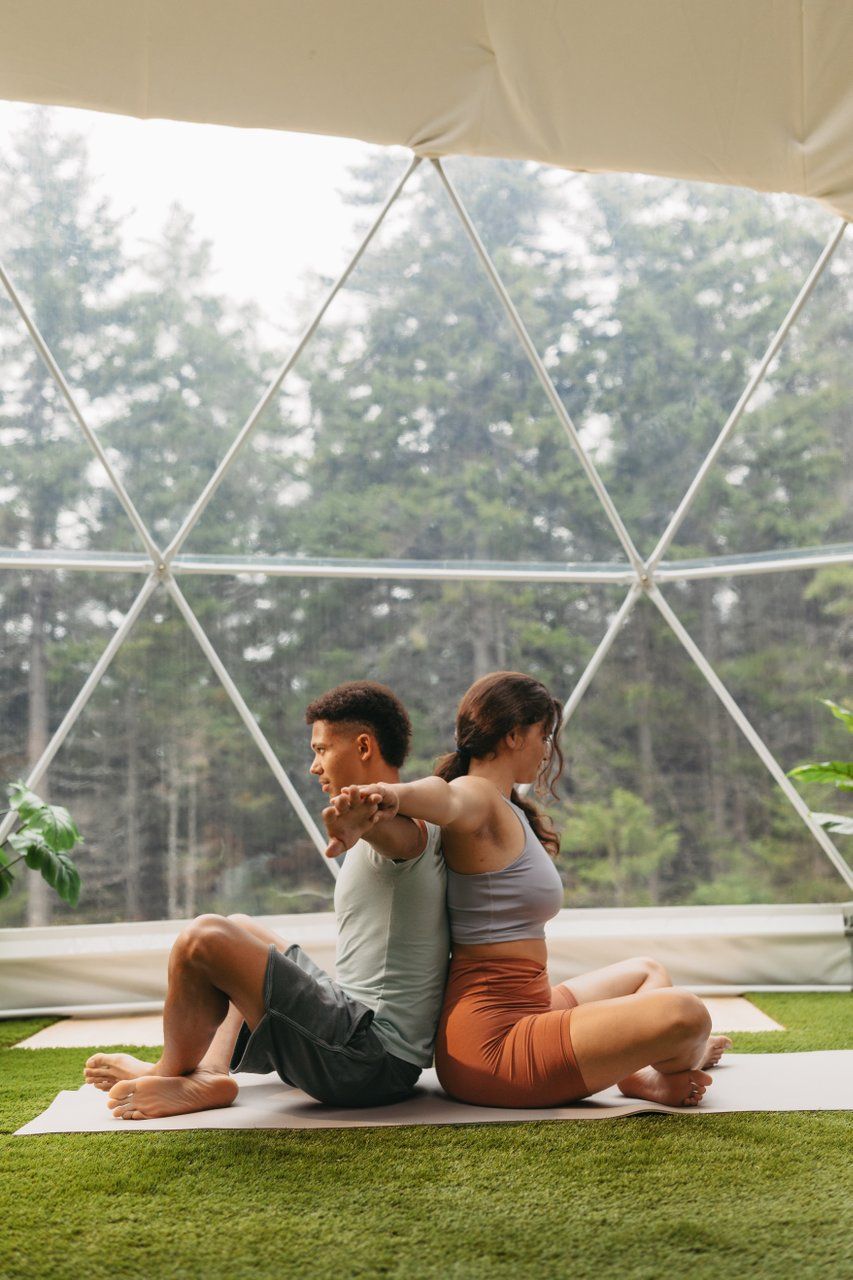Twee mensen zitten met hun ruggen naar elkaar toe op yogamatten in een tent en doen rek- en strekoefeningen in het gras in de buitenlucht.